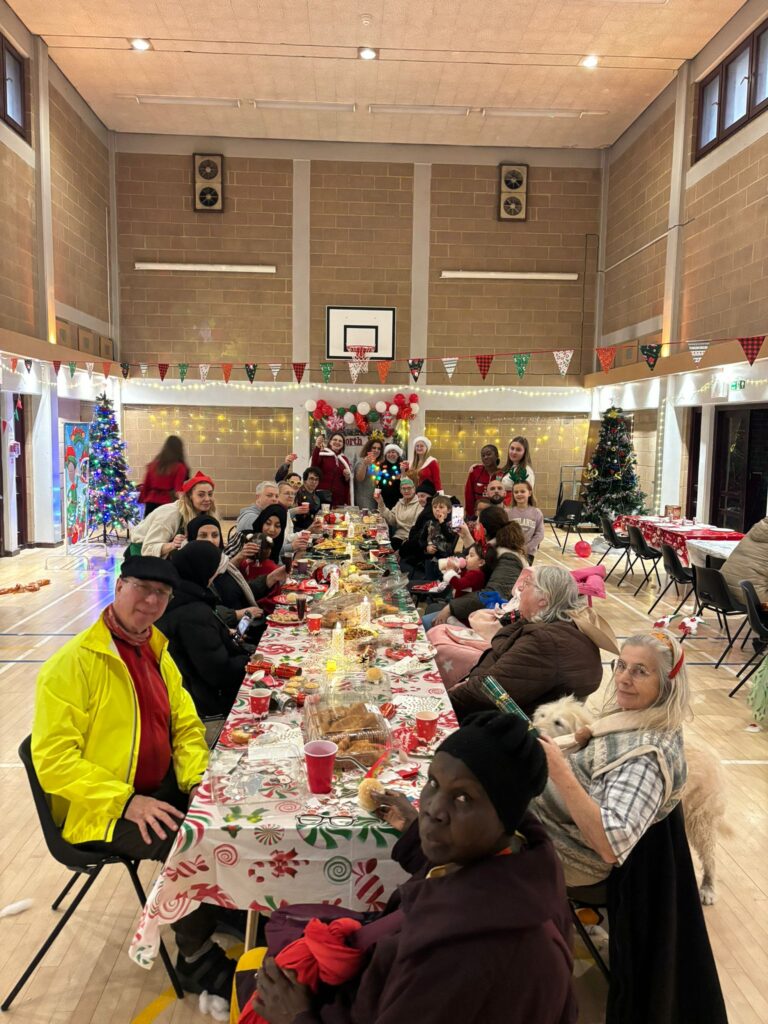 A community group sit on a long table in a sports hall and enjoy a Christmas lunch together