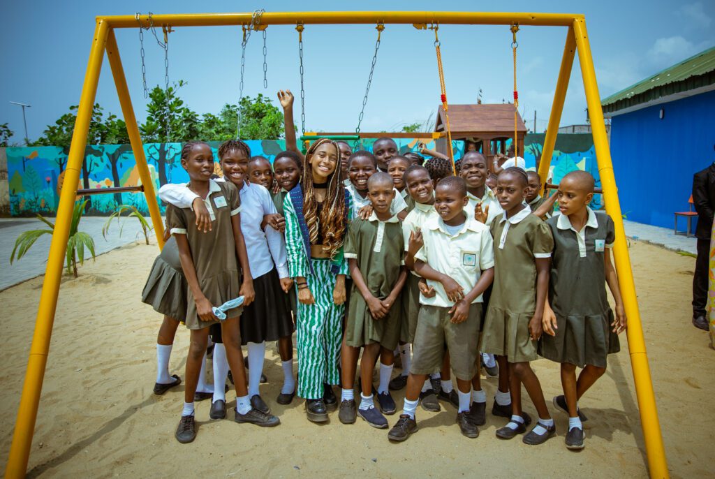 A large group of happy children gather under a set of yellow swings, with the architect and project leader, 17 year old Amara, stood infront