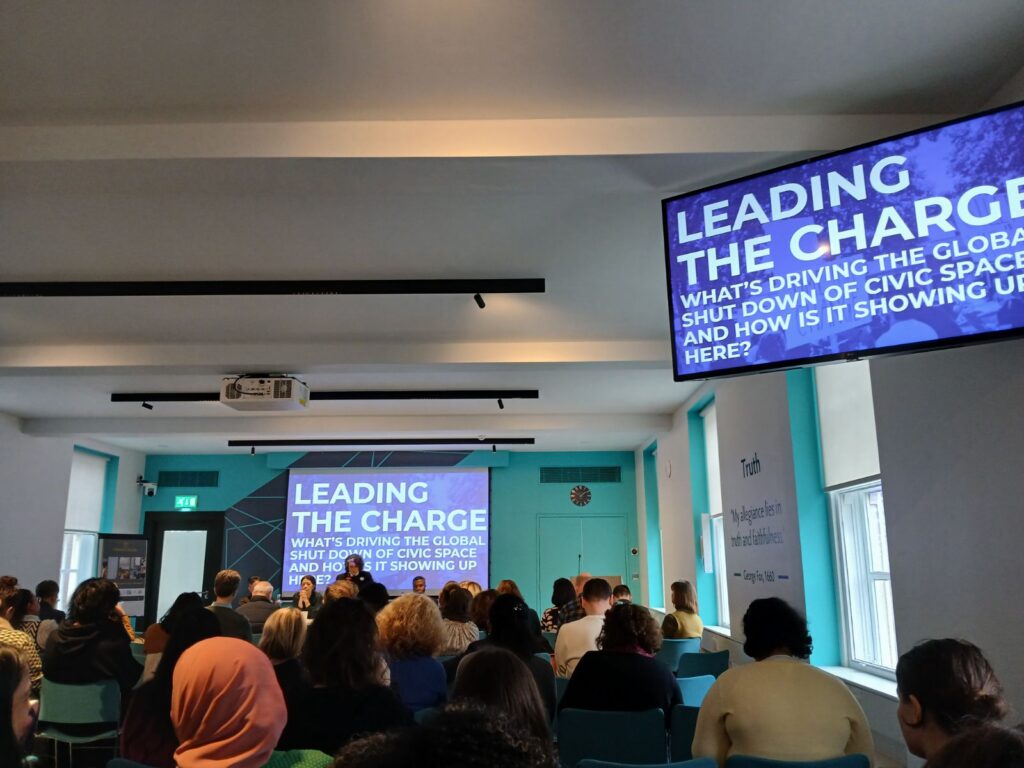 In the room at the Speak Out event, a large projected screen is infront of a busy seated audience
