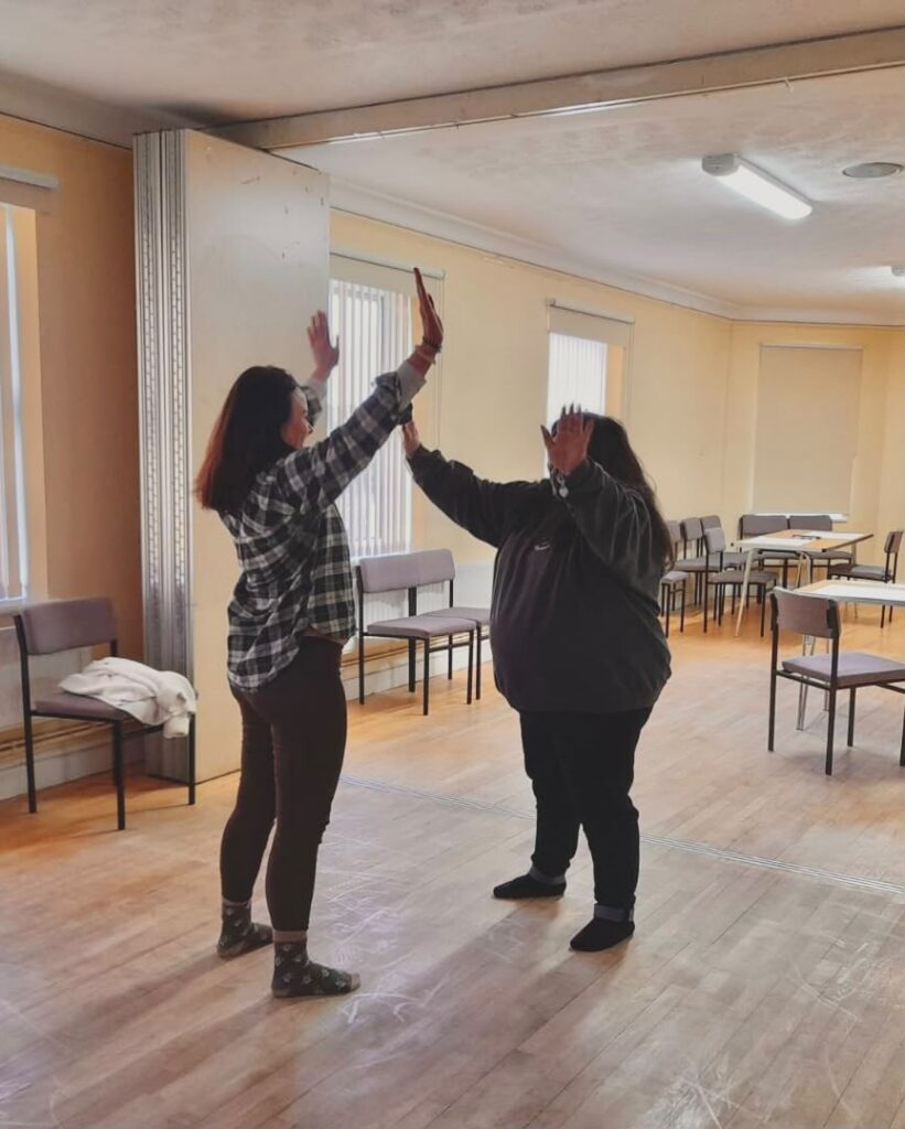Two women in a room doing a mirroring exercise, both with their arms above their heads